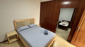 Un lit simple avec une tête de lit en bois et une petite table de chevet, dans une pièce qui comprend également une armoire en bois et une porte donnant sur une autre pièce.