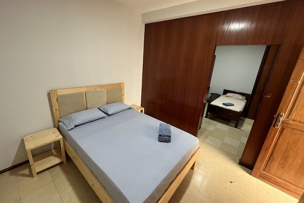 A single bed with a wooden headboard and a small bedside table in a room with a wooden wardrobe and a door leading to another room.