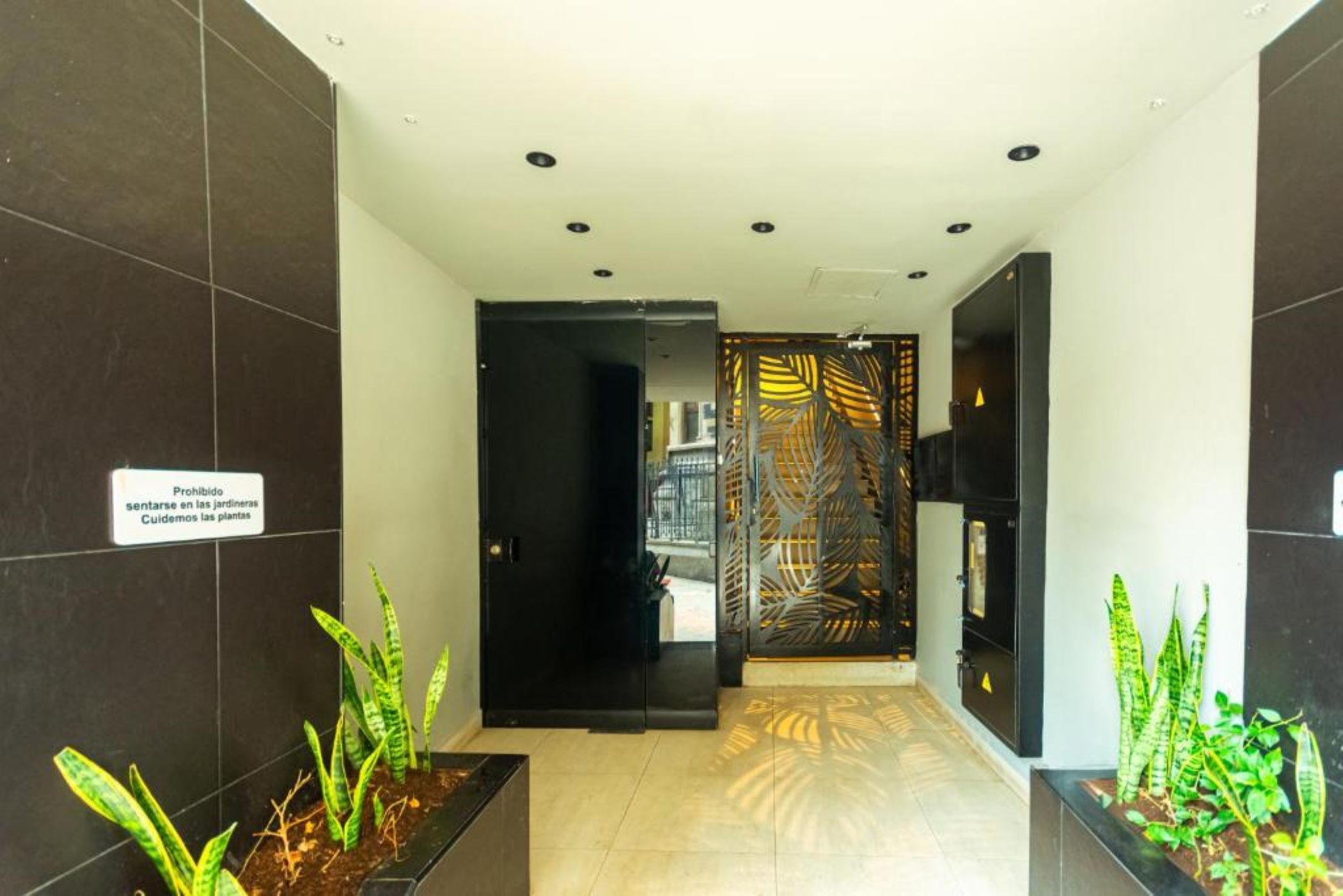A hallway with dark tiled walls, a sign prohibiting sitting in the gardens, and potted plants at the end.