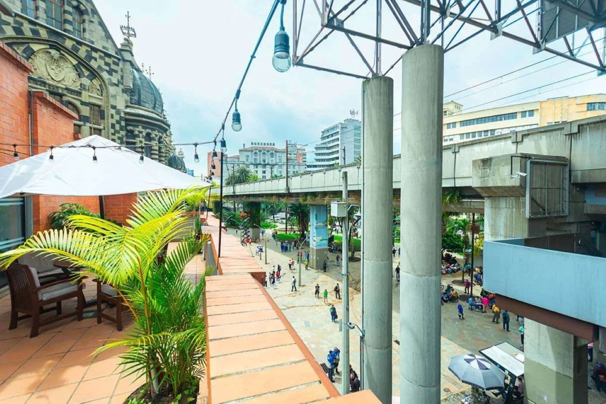 An outdoor urban space with a covered seating area, a palm tree, and a pedestrian walkway.