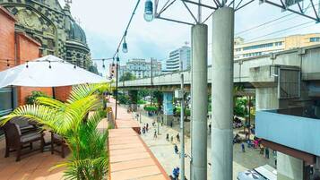 An outdoor urban space with a covered seating area, a palm tree, and a pedestrian walkway.