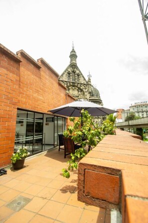 A patio area with a brick wall, a glass door, a table with an umbrella, and a building with a clock tower in the background. - Mandala Medellin Botero By Gyc Hotels (Medellín)