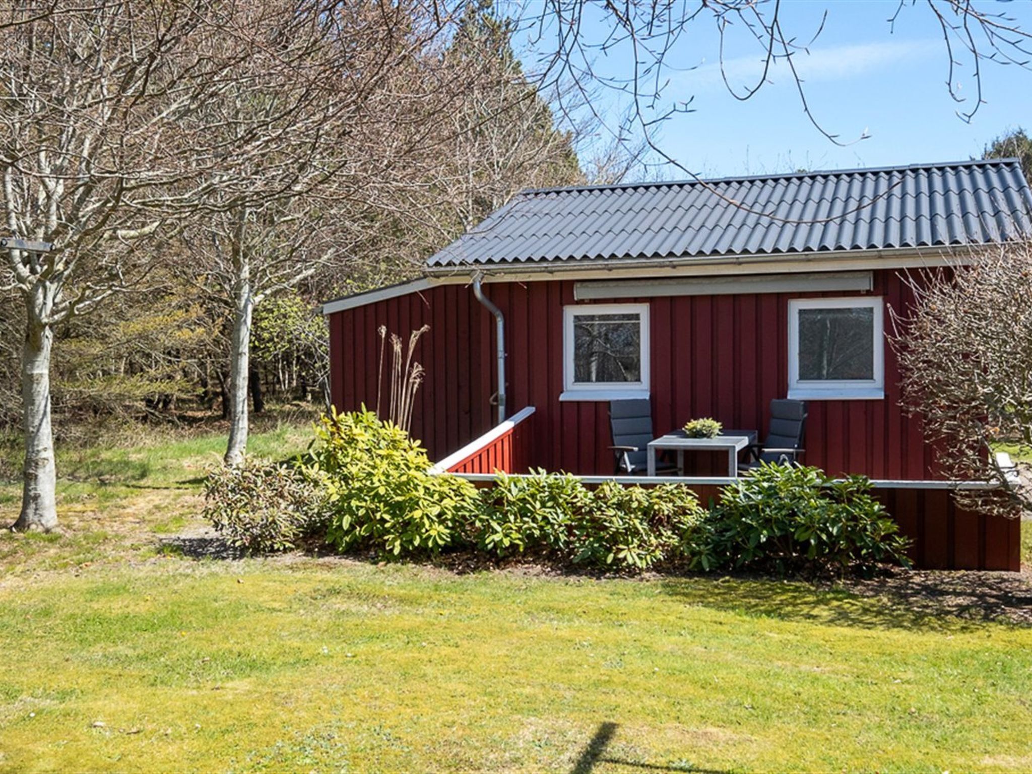 Een rode houten hut met een metalen dak, twee ramen en een klein terras met een tafel en stoelen.
