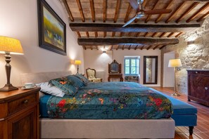 Quarto com cama, cabeceira de madeira, ventilador de teto, parede de pedra e janela.
