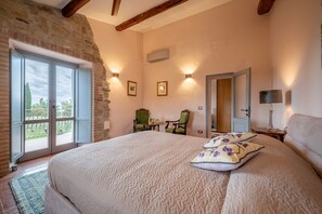A bedroom with a large bed, a chair, a table, a lamp, and a door leading to another room.