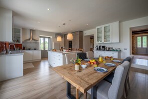 A modern kitchen with a central island, wooden dining table, and a view of the outdoors. - Tenuta Santa Cristina 10, Emma Villas (Gubbio)