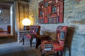 Um quarto com paredes de pedra, duas poltronas estampadas, uma pequena mesa de madeira e uma estante fixada na parede.