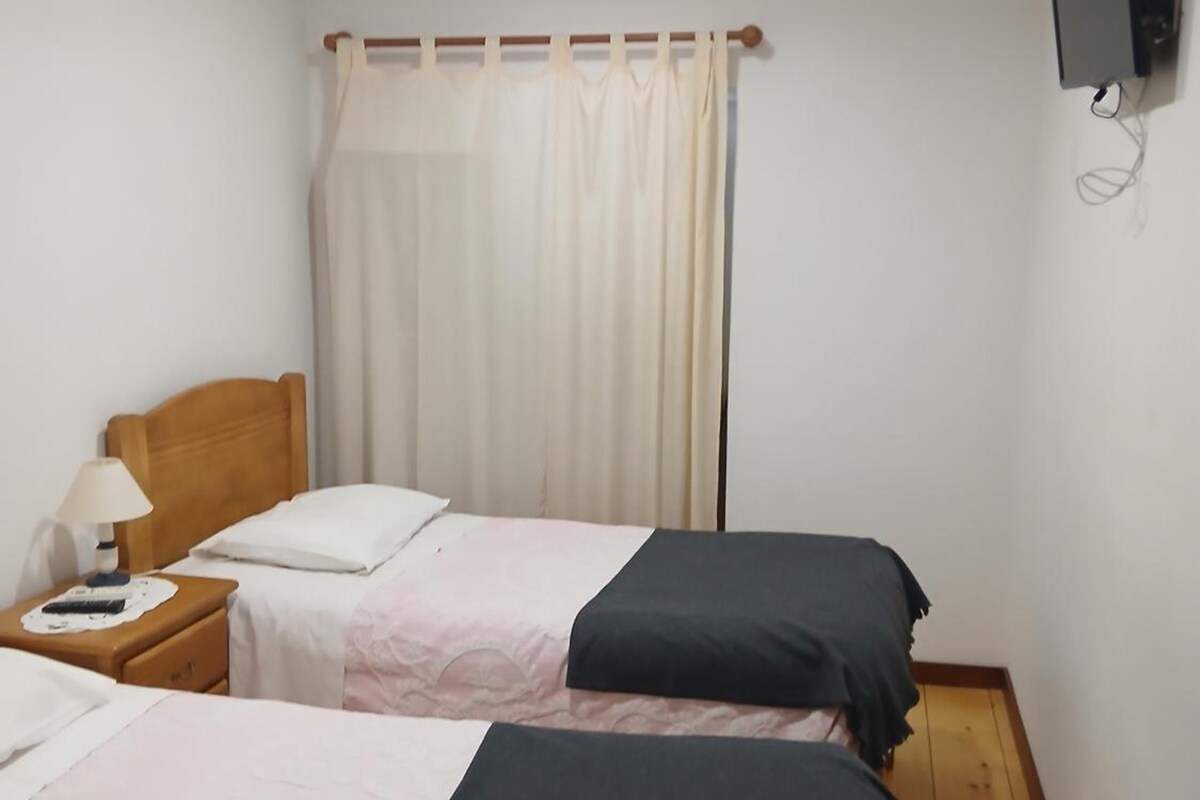 A room with two beds, a nightstand with a lamp, a curtain, and a television mounted on the wall.