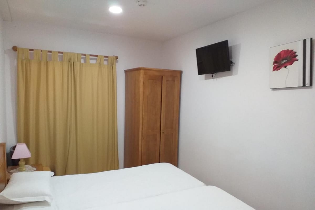 Un dormitorio con una cama, un armario de madera, un televisor en la pared y una ventana con cortinas.