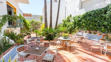 Un patio al aire libre con varias zonas de estar, incluyendo mesas y sillas, rodeado de plantas diversas y un suelo de baldosas.