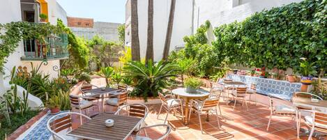 Un patio al aire libre con varias zonas de estar, incluyendo mesas y sillas, rodeado de plantas diversas y un suelo de baldosas.