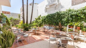 Un patio al aire libre con varias mesas y sillas, rodeado de plantas en macetas y una pared blanca.