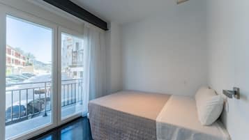 Una habitación pequeña con cama individual, balcón, paredes blancas y vista a una calle y edificios.