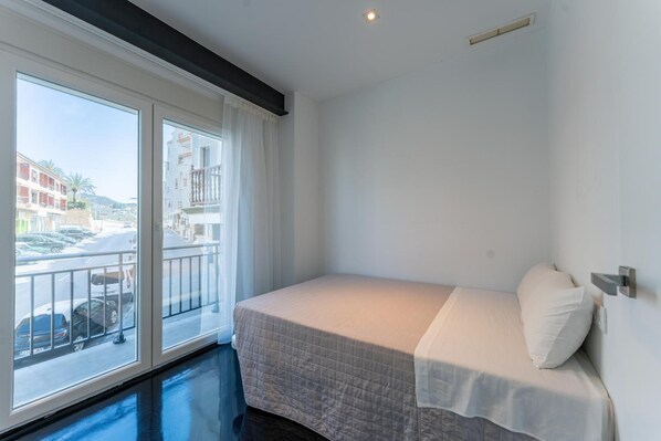 A small, single-bed room with a balcony, white walls, and a view of a street and buildings. - Hotel Buigues (Teulada)