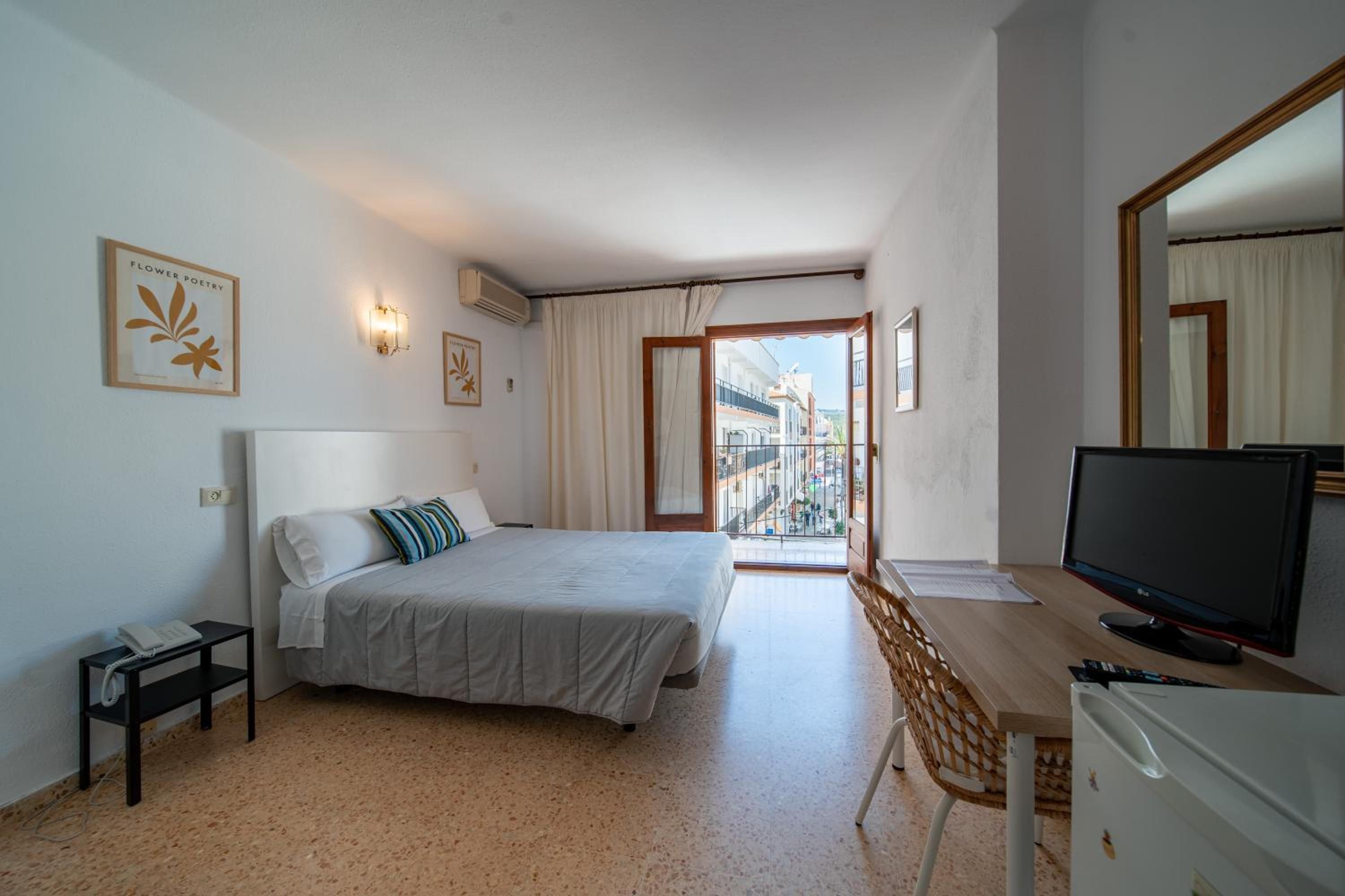 Habitación de hotel con una cama, un escritorio con televisor, una silla y vistas de la ciudad desde un balcón.