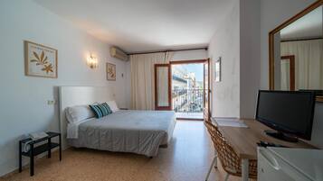 Habitación de hotel con una cama, un escritorio con televisor, una silla y vistas de la ciudad desde un balcón.