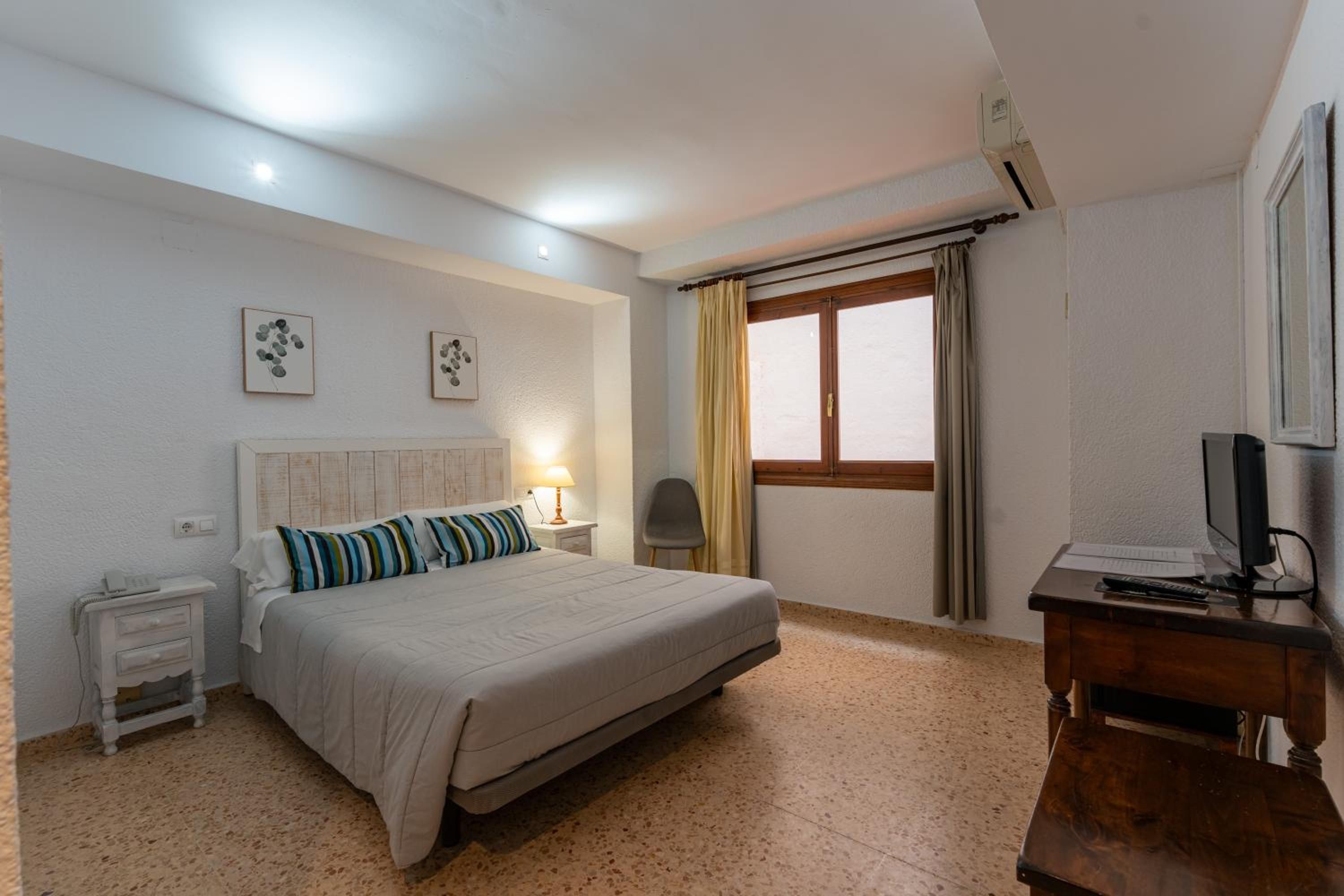 Habitación de hotel con cama, mesitas de noche, un escritorio con computadora, una silla, ventana con cortinas y decoración en la pared.