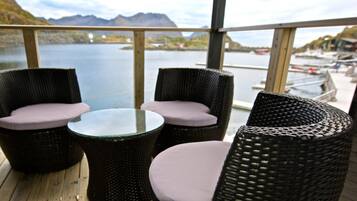 A deck with wicker furniture and a glass table, overlooking a lake with boats and mountains in the distance.