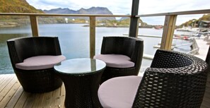 A deck with wicker furniture and a glass table, overlooking a lake with boats and mountains in the distance.