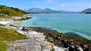 A rocky shoreline with clear turquoise water, a person sitting on the rocks, and mountains in the background.