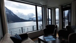 Habitación con una ventana grande que da a un lago y montañas, un sofá, dos sillones y una mesa con papeles.