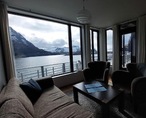 A room with a large window overlooking a lake and mountains, a sofa, two chairs, and a table with papers. - Hamn I Senja (Senja)