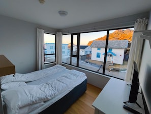 A bedroom with a bed, a desk, and a large window with a view of buildings and a mountain. - Hamn I Senja (Senja)