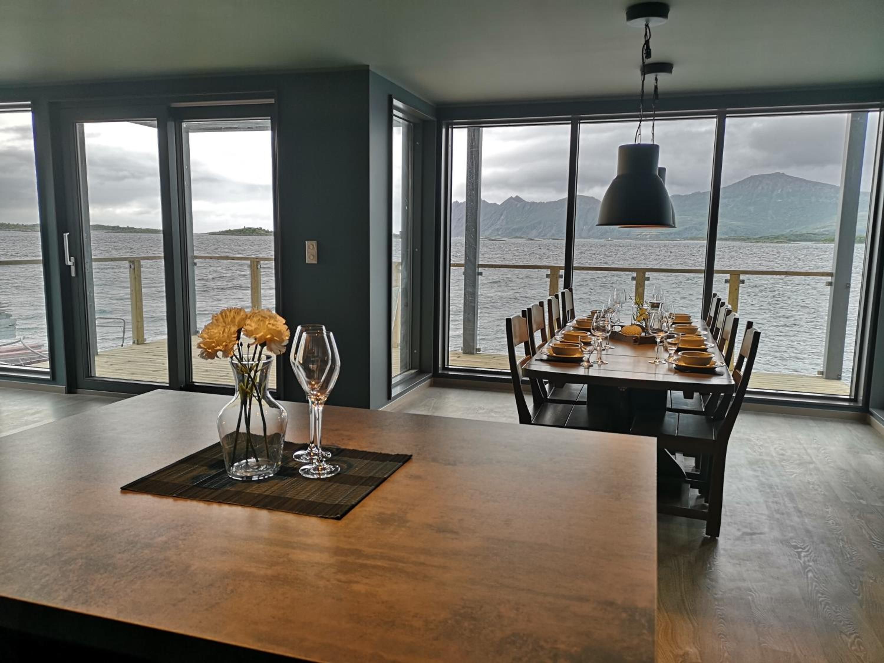 Comedor con una mesa dispuesta para seis personas, ventanas amplias con vistas al mar y a las montañas, y una lámpara colgante.