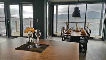 Comedor con una mesa dispuesta para seis personas, ventanas amplias con vistas al mar y a las montañas, y una lámpara colgante.