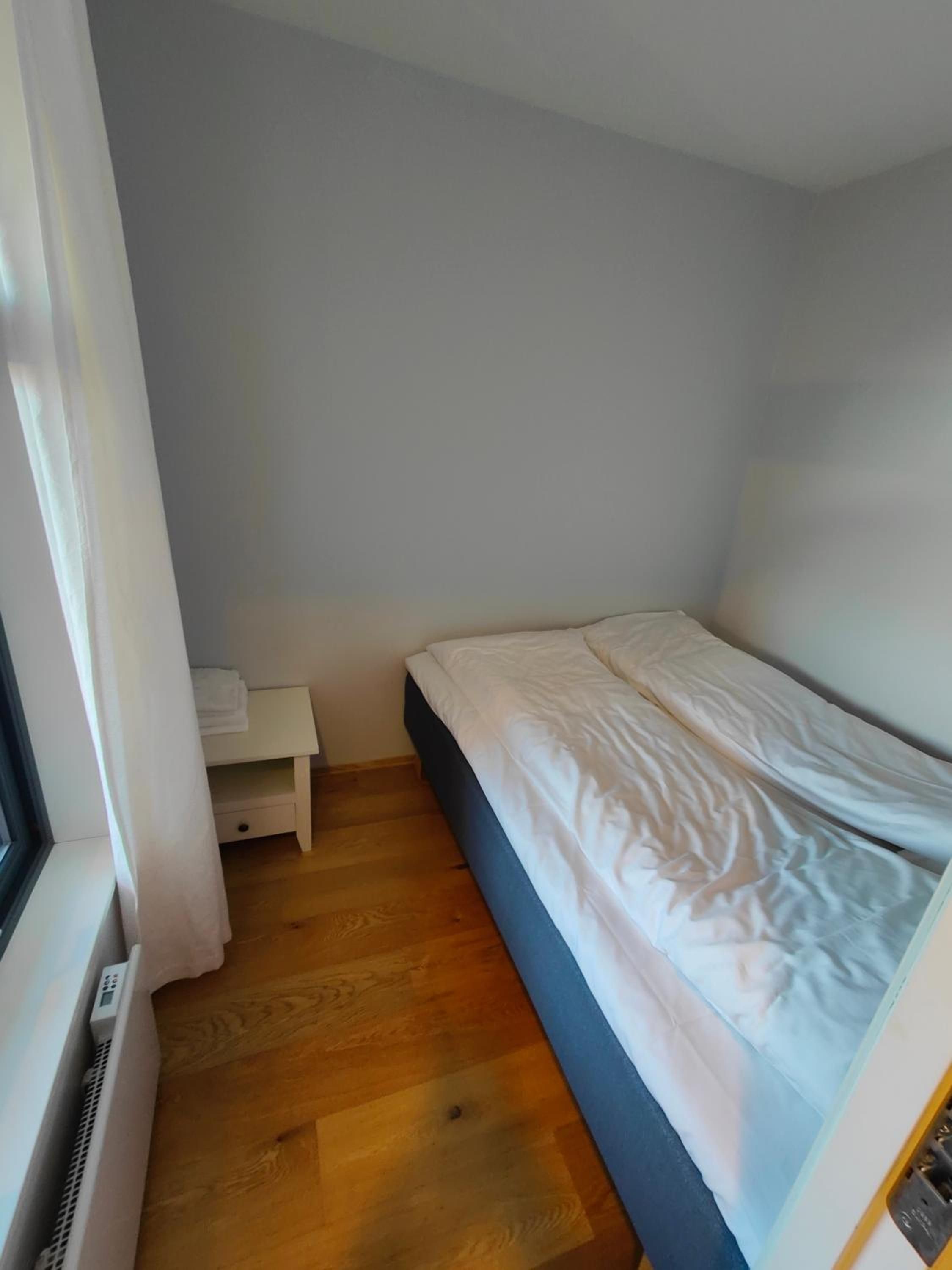 A small, single-bed room with a wooden floor, a white bedside table, and a window.