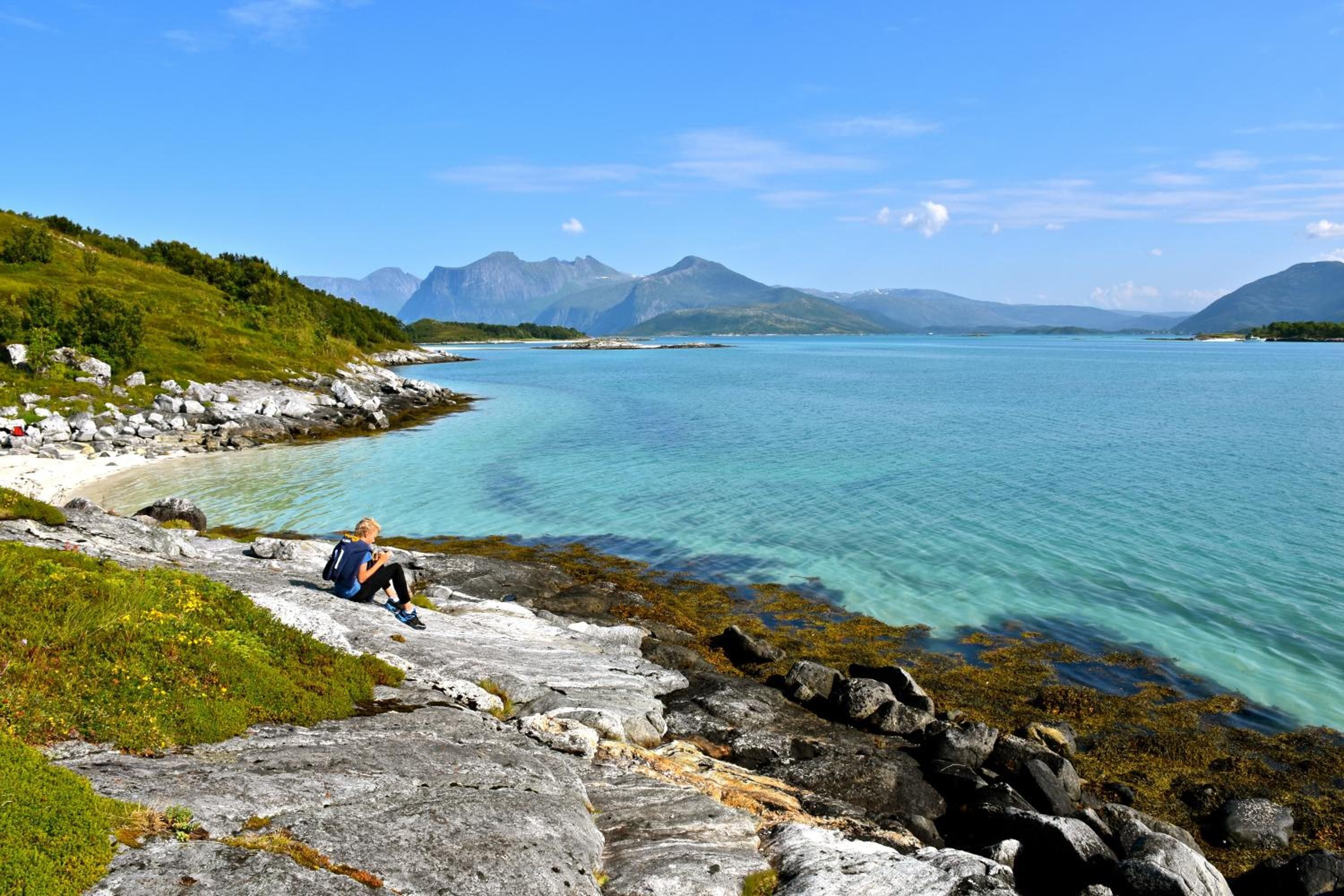 A rocky shoreline with clear turquoise water, a person sitting on the rocks, and mountains in the background.