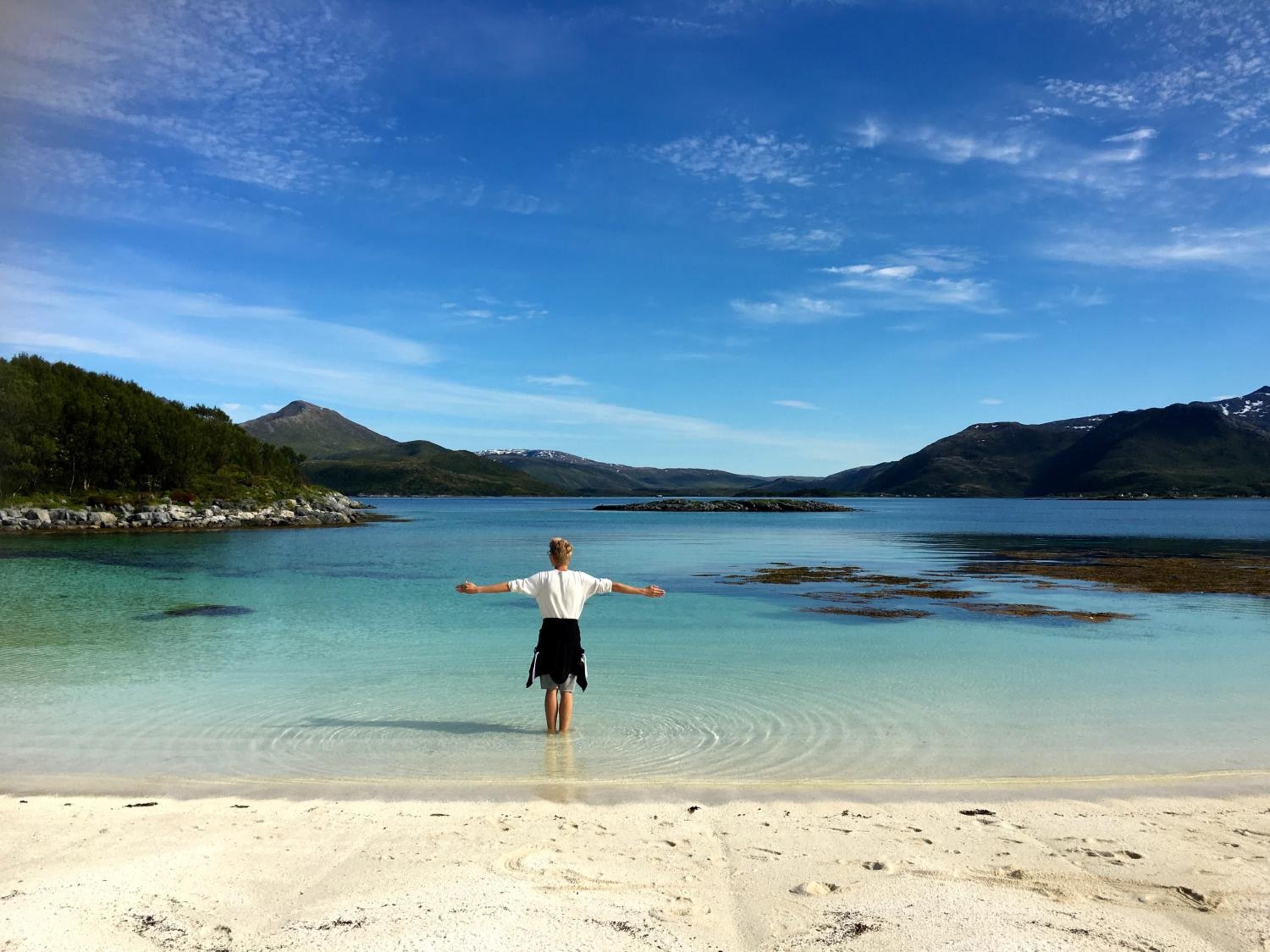 A person standing on a beach with arms outstretched, facing a calm lake with mountains in the background.