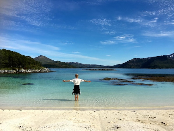 A person standing on a beach with arms outstretched, facing a calm lake with mountains in the background.