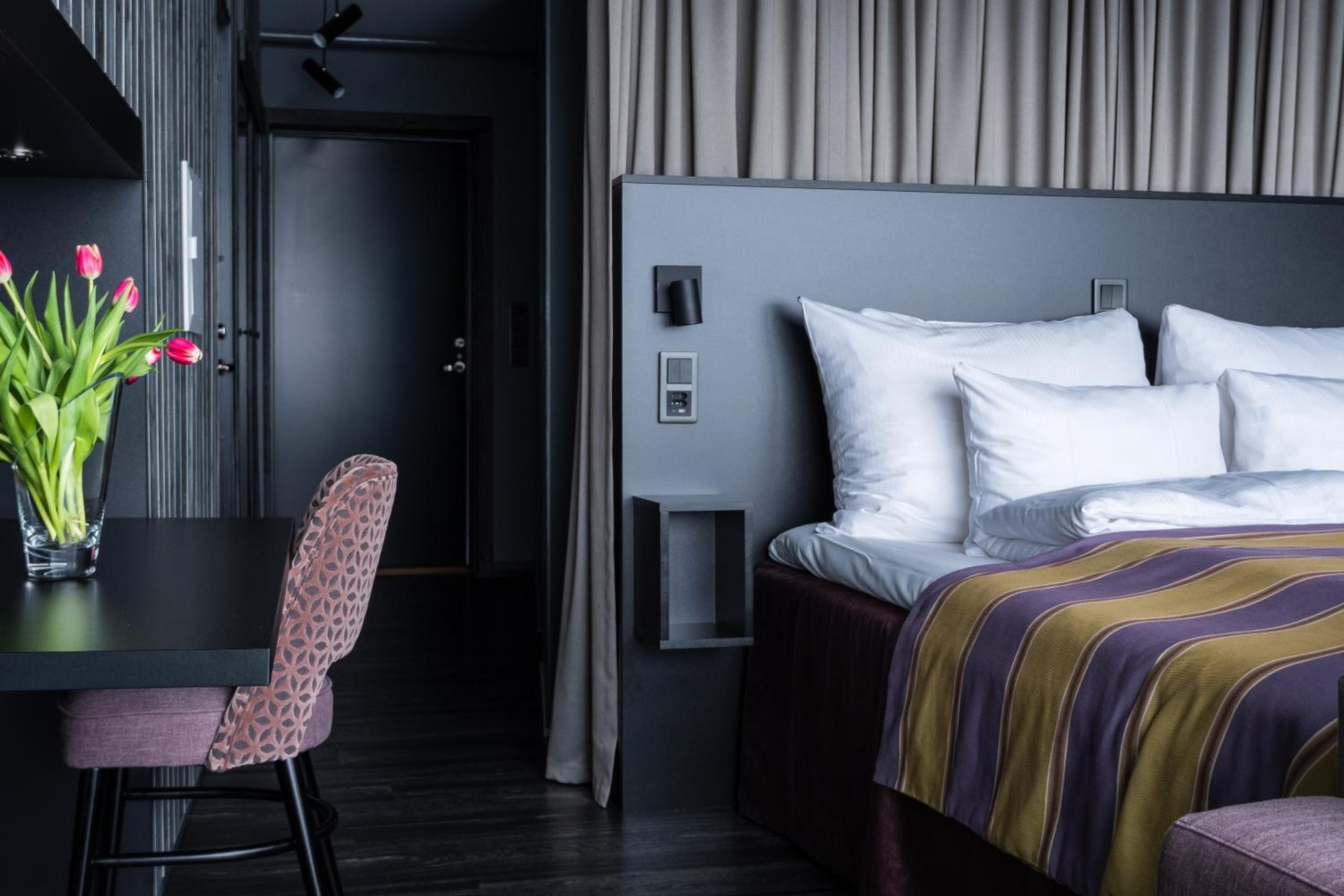 Une chambre d’hôtel moderne, équipée d’un lit, d’un bureau avec une chaise et d’un vase de fleurs.