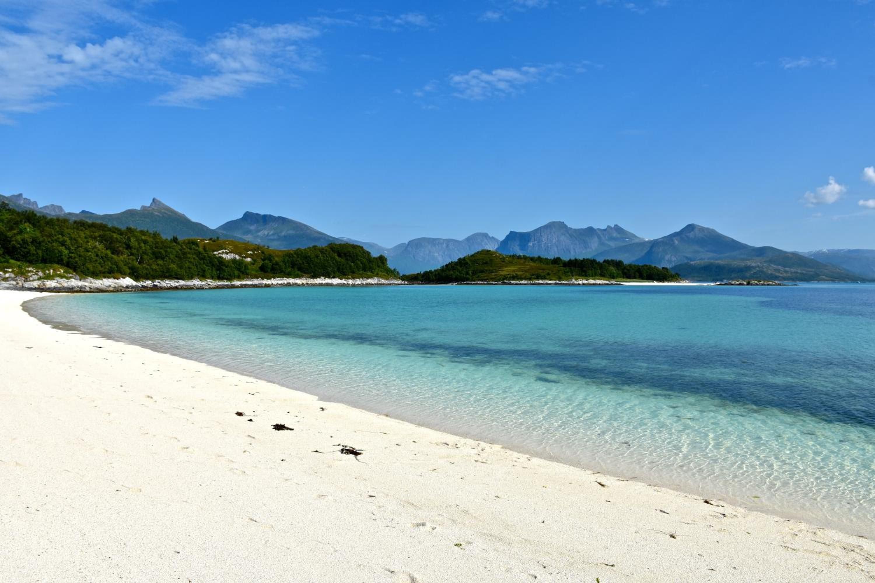 A beach with clear turquoise water, white sand, and a forested island with mountains in the background.