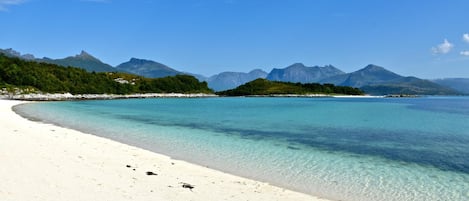 A beach with clear turquoise water, white sand, and a forested island with mountains in the background.