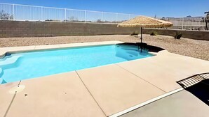 Ein Schwimmbecken mit Betonumrandung, weißem Zaun und einem reetgedeckten Gebäude im Hintergrund.