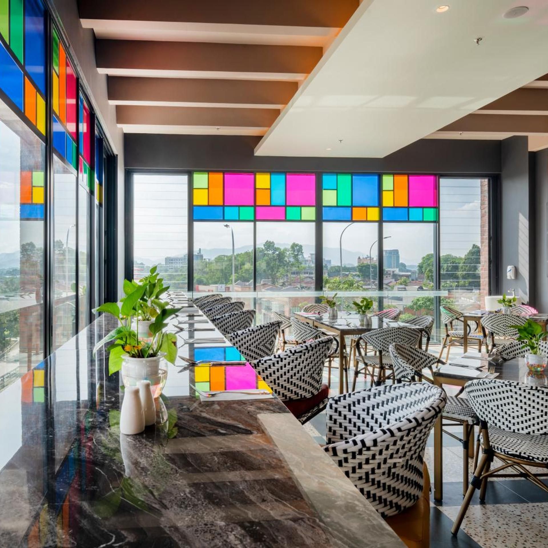 A modern interior with a long dining table, wicker chairs, and large windows offering a city view.