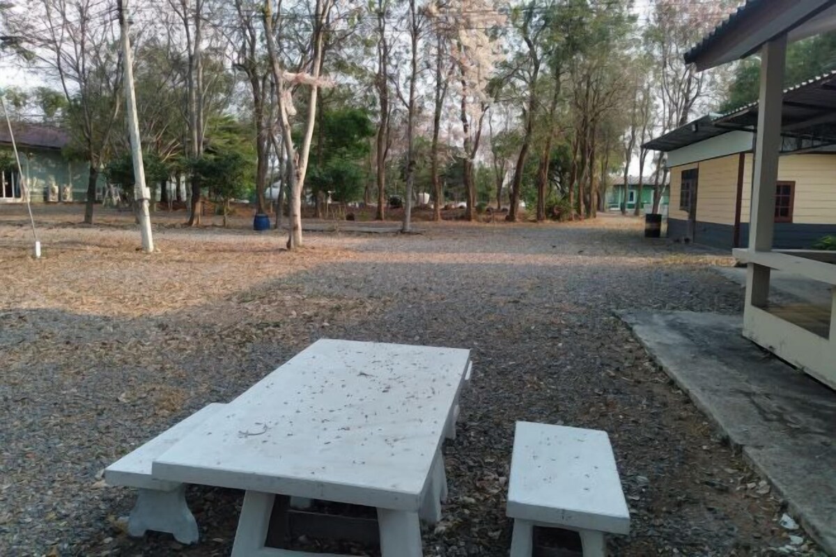 An outdoor seating area with two white tables and benches, surrounded by trees and a building with a covered porch.
