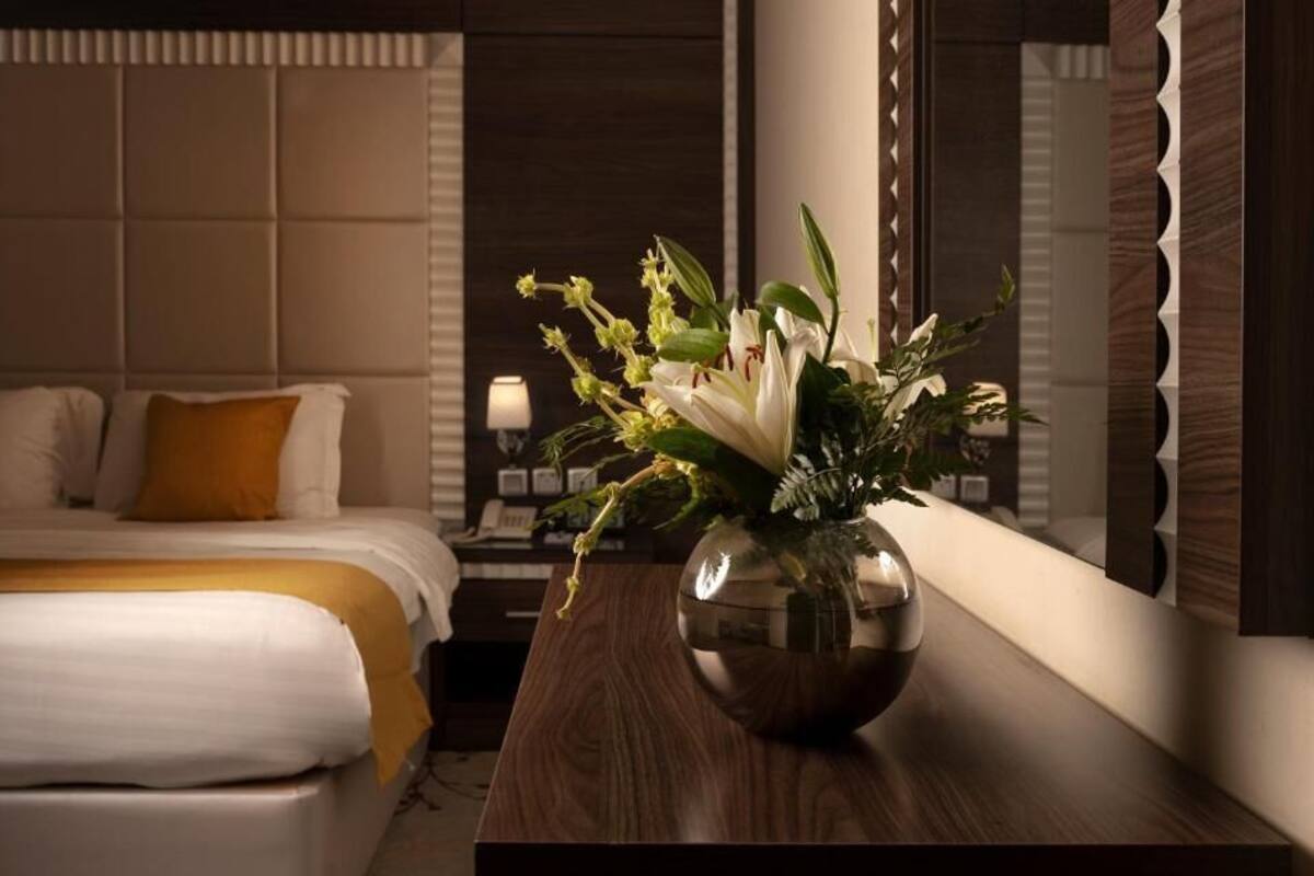 A hotel room with a bed, a nightstand, a vase with flowers, and a large mirror.