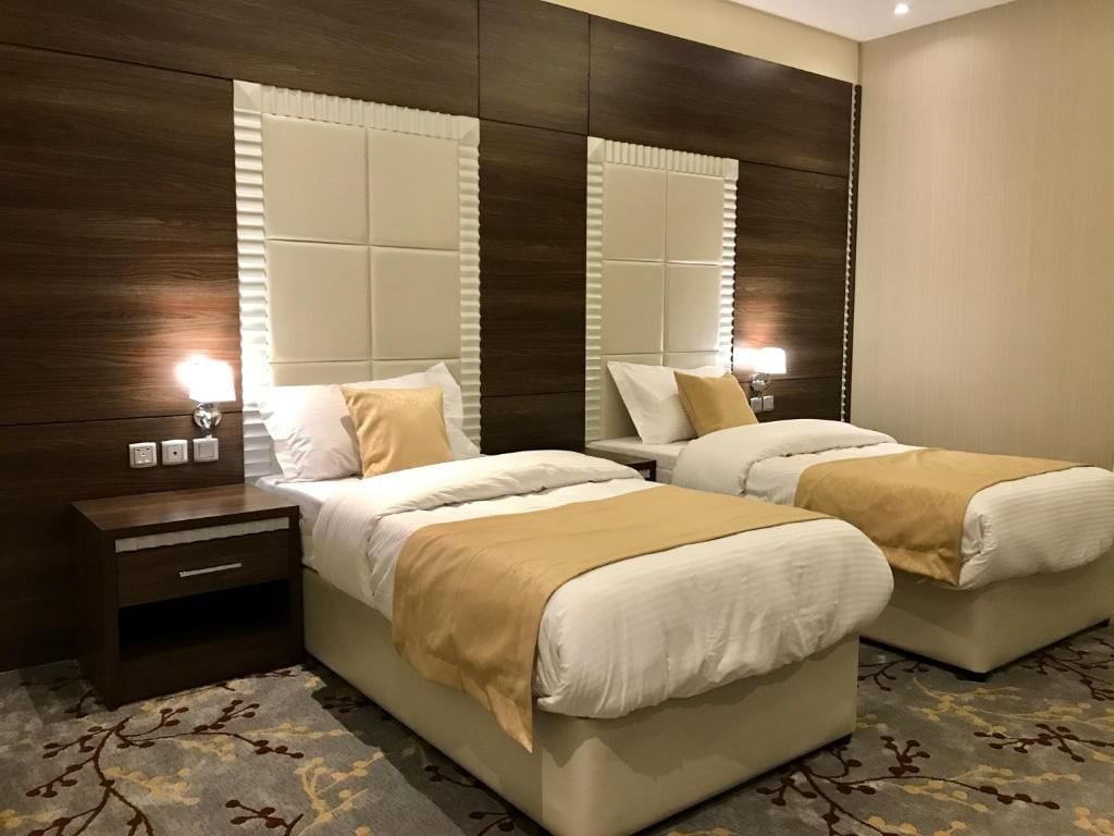Habitación de hotel con dos camas, cabecero de madera, mesita de noche y alfombra estampada.