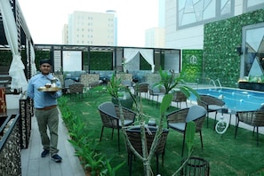 Um bar no terraço com móveis de vime, piscina e vista para prédios.