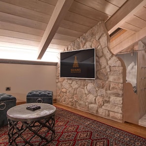 A cozy room with a stone wall, a mounted television, a round table with a remote, and two cushioned seating areas. - Kamares Boutique Hotel & Spa Ioannina (Ioannina)
