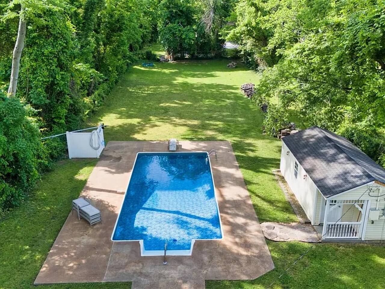 Et rektangulært swimmingpool med betondæk, omgivet af græsplæne og frodige træer.