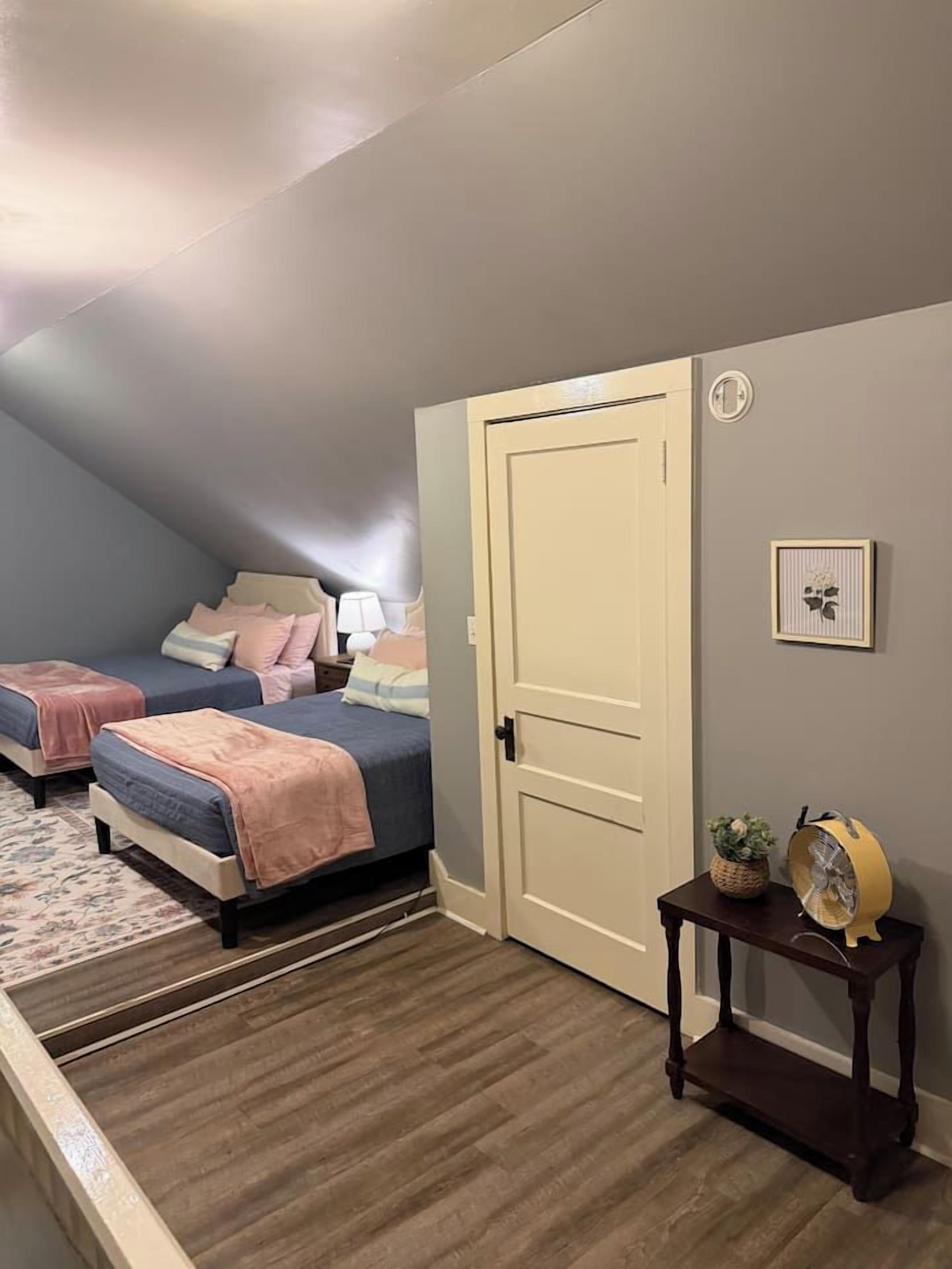 Ein Schlafzimmer mit zwei Betten, einer Tür, einem kleinen Tisch mit Ventilator und einem gerahmten Bild an der Wand.