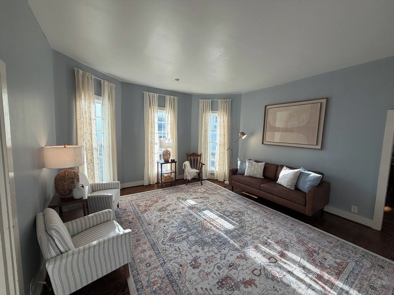 A spacious living room with a large patterned rug, a brown sofa, striped armchairs, a wooden side table with a lamp, and a chair.