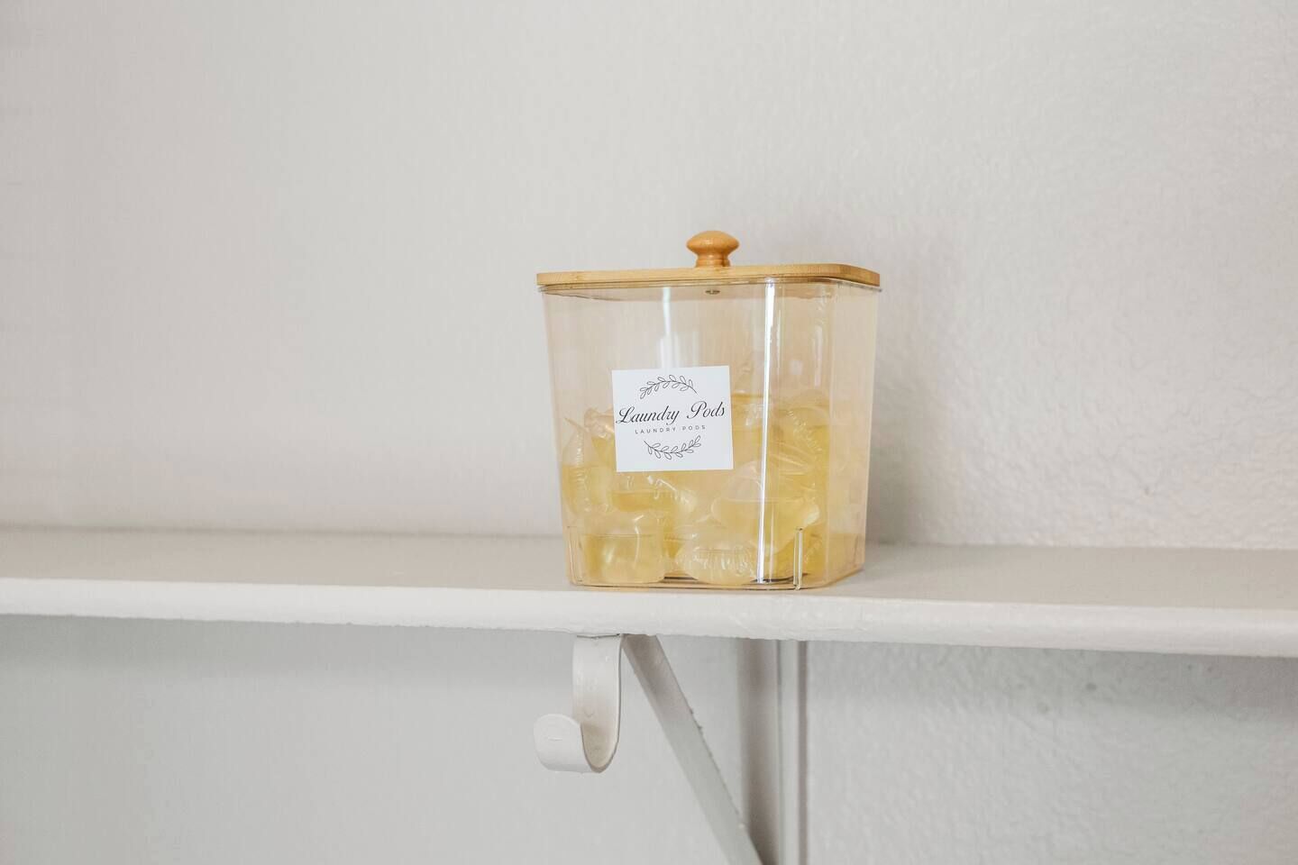 Un récipient transparent en verre, doté d’un couvercle en bois, sur lequel est inscrit « Dosettes de lessive », et qui est posé sur une étagère blanche.