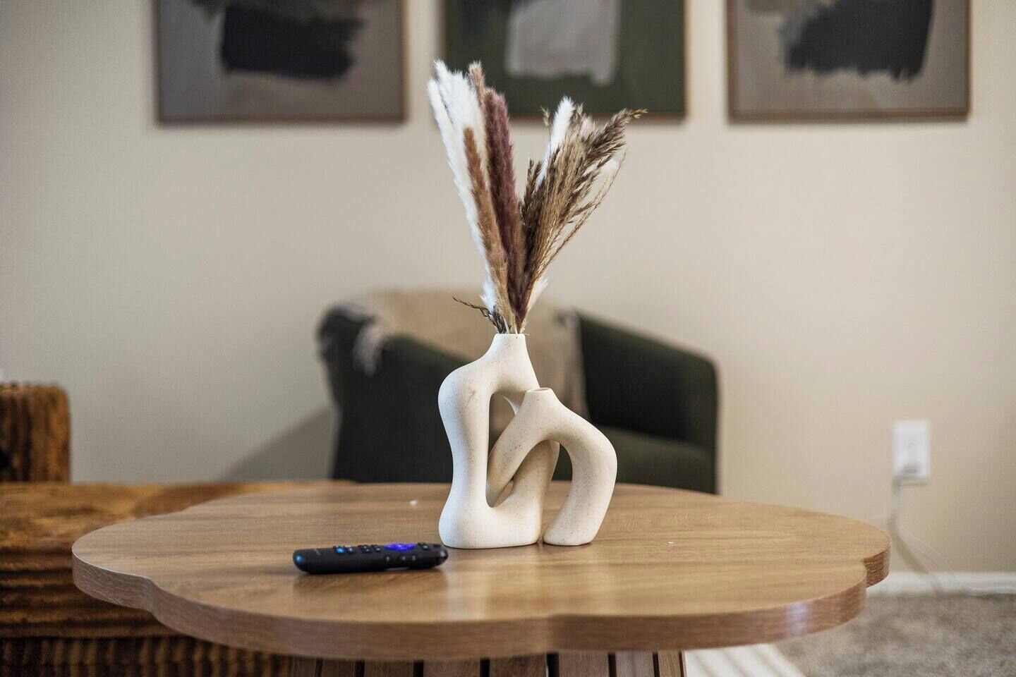 A round wooden table with a vase containing dried pampas grass and a remote control.