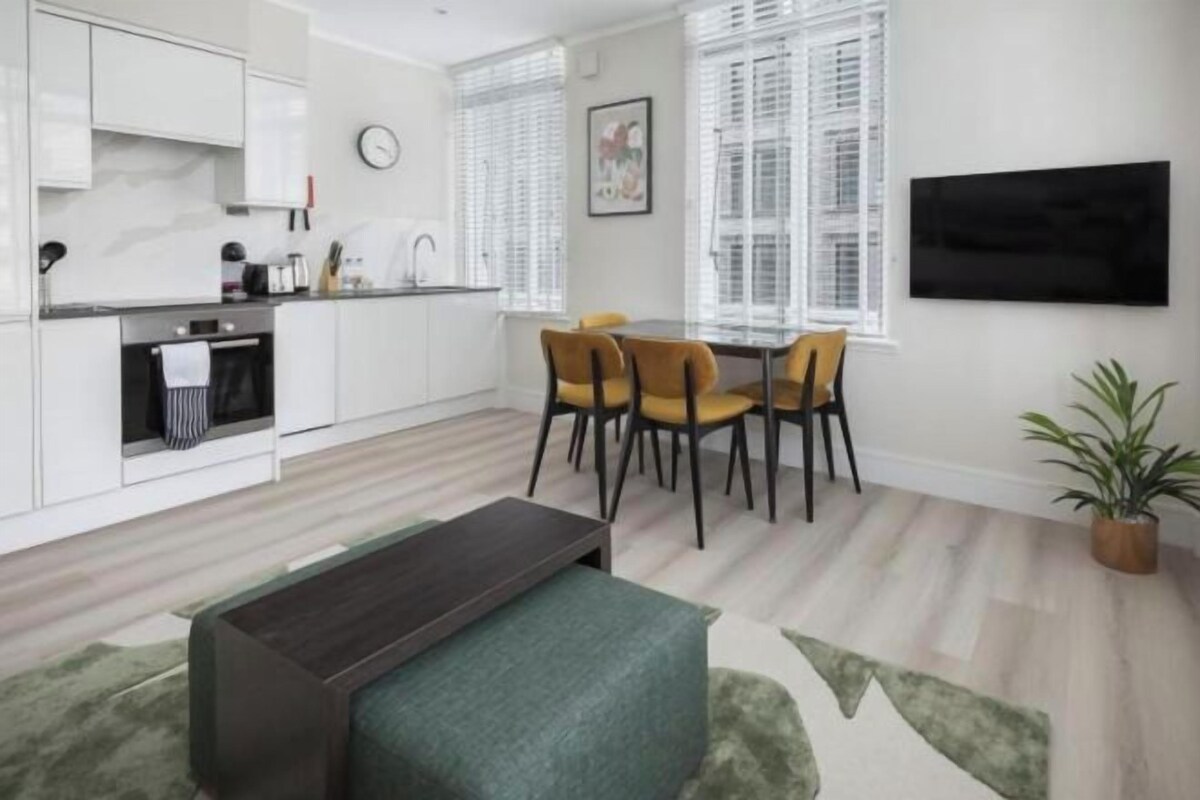 Eine moderne Küche mit weißen Schränken, ein Essbereich mit Tisch und Stühlen sowie ein an der Wand befestigter Flachbildfernseher.
