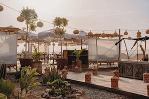 An outdoor patio area with tables and chairs, covered by umbrellas, and surrounded by potted plants and decorative lanterns. - Mia Agafay Resort (Agafay)